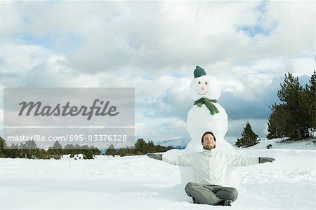 Young man sitting on ground in front of snowman, smiling, arms outstretched