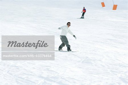 Young man snowboarding on ski slope, full length, skier in background