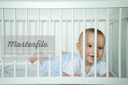 Baby lying on stomach, looking through bars of crib, smiling