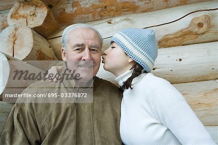 Senior man smiling at camera, granddaughter kissing his cheek, portrait