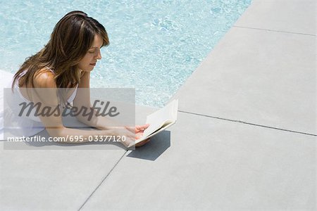 Woman lying on stomach next to pool, reading book