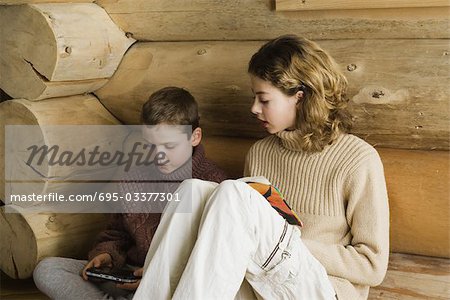 Teen girl and preteen boy sitting side by side, reading and playing video game