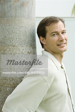 Man leaning against tree trunk, smiling over shoulder at camera