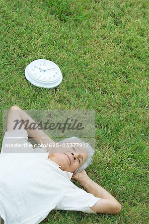 Senior man lying on grass next to clock, hands behind head, high angle view