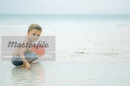 Little boy crouching in water at the beach, playing with beach ball, looking at camera