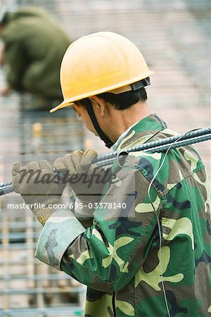 Construction worker wearing camouflage and hard hat carrying metal rod on shoulder, side view