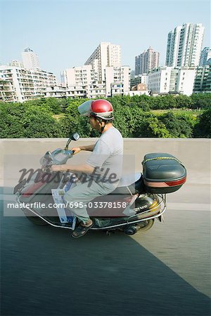 China, Guangzhou, man riding motor scooter, high-rise buildings in background, side view