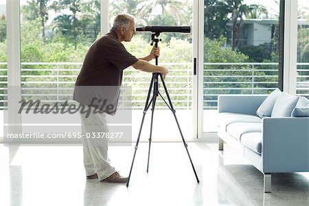 Man standing in living room, looking through telescope, side view