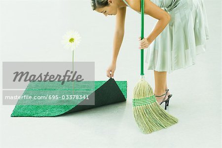 Woman bending over, broom in hand, lifting corner of artificial turf next to gerbera daisy