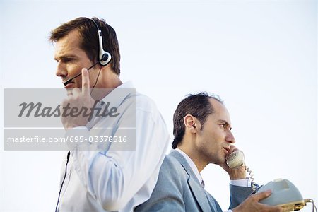 Two businessmen back to back, one using headset, the other using landline phone, side view