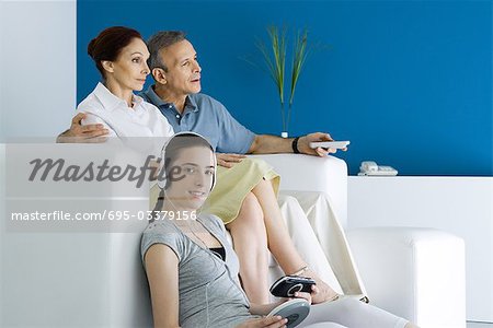Teen girl listening to portable CD player, smiling at camera, her parents watching TV in background