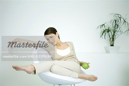 Young woman slouching in chair, holding apple, smiling at camera