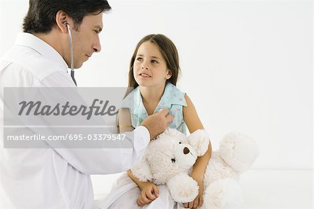 Pediatrician listening to girl's heart with stethoscope, girl holding teddy bear