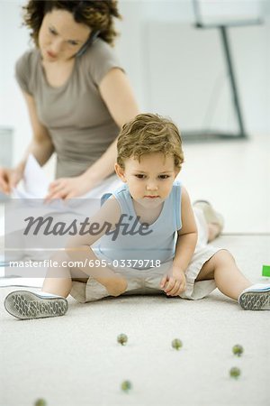 Boy playing with marbles while young mother uses phone and looks at paperwork in background