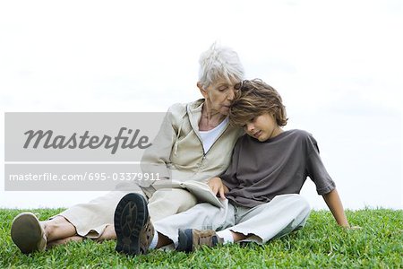 Grandmother and grandson sitting side by side on the ground, reading book together