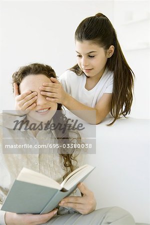 Girl standing with hands over her mother's eyes, woman holding book
