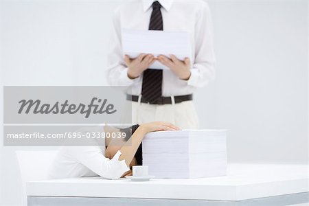 Woman with head down on desk, next to tall stack of papers, man holding another stack of paper