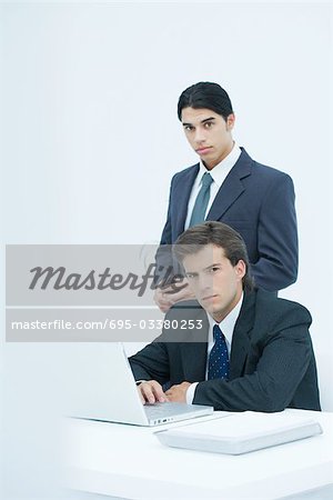 Two serious businessmen looking at camera, one sitting at desk, one standing behind him