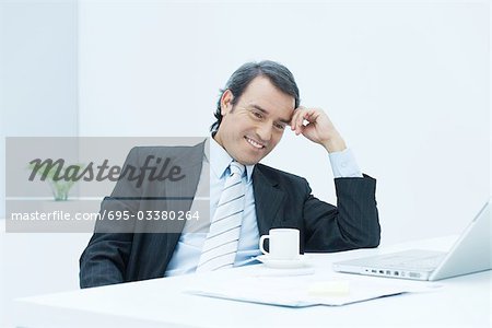 Businessman sitting at desk, leaning on elbow and holding head, looking down and smiling