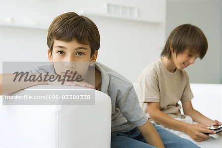 Boys sitting on couch, one looking at camera, the other playing handheld video game