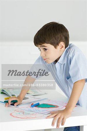 Boy leaning on table, holding crayon, colorful drawing on paper and the table