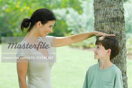 Boy standing against tree trunk, his mother placing her hand on his head, both smiling looking at each other