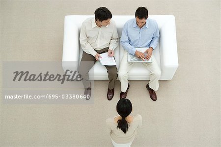 Professional men sitting on sofa, discussing document, woman standing before them, overhead view