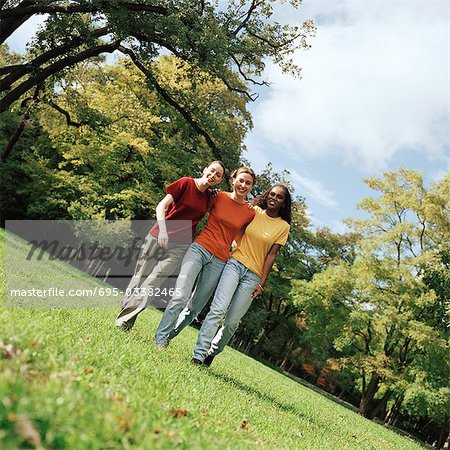 Three young women walking on grass with arms around each other, front view, full length