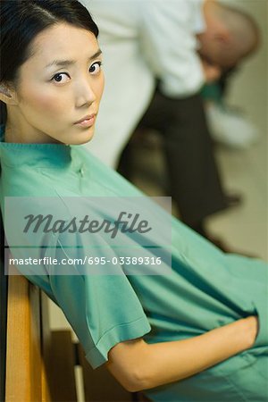 Female medical worker sitting with hand in pocket, looking at camera, doctor with head down in background