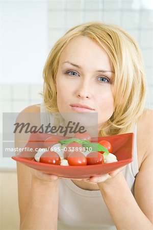 Woman holding up a plate of Caprese salad, looking at camera