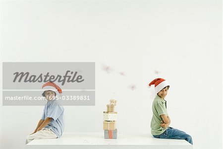 Two brothers sitting back to back, Christmas gifts stacked between them, both wearing Santa hats