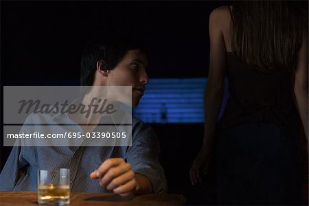Glass of whiskey on table, man sitting and looking away, rear view of woman standing in the dark