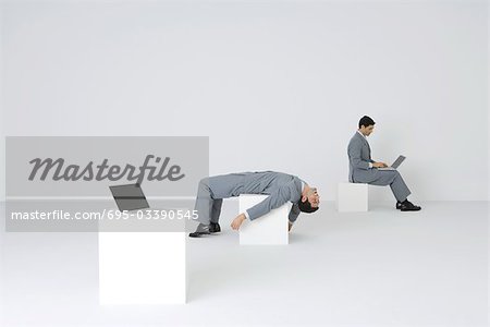 Identical businessmen, one using laptop while the other lies on back, exhausted