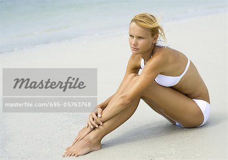 Woman in bikini sitting on beach, hugging knees, full length