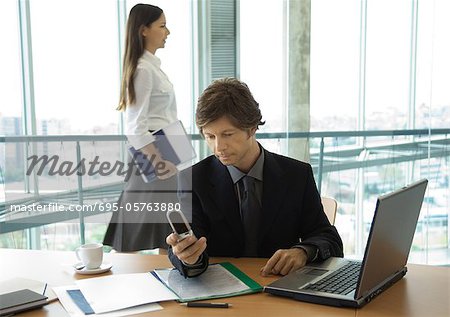 Businessman looking at cell phone, woman walking by in background