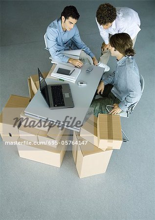 Three male colleagues grouped around cell phone on desk, surrounded by cardboard boxes