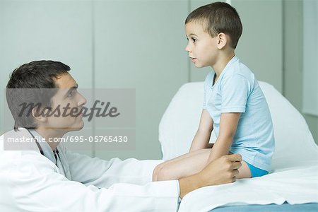 Boy sitting on examination table, doctor looking up at boy, profile