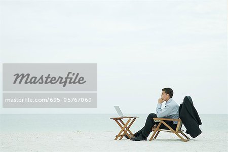 Businessman sitting on beach, looking at laptop computer, hand under chin, full length