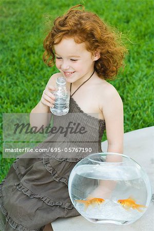 Girl sitting next to goldfish bowl, drinking water