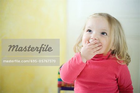 Blonde toddler girl covering mouth, looking up