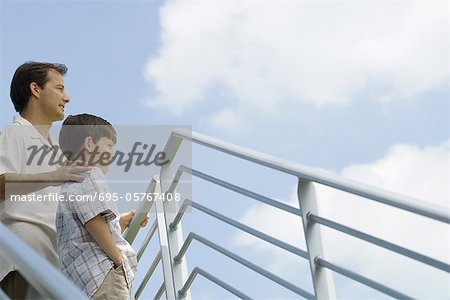 Father and son standing by railing looking at sky, low angle view