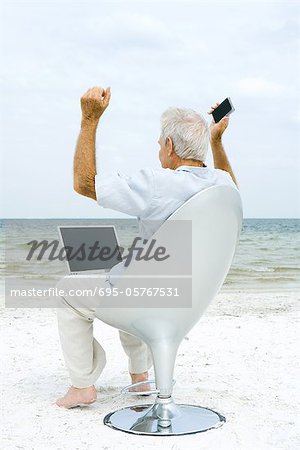 Senior man using laptop and cell phone on beach, raising arms