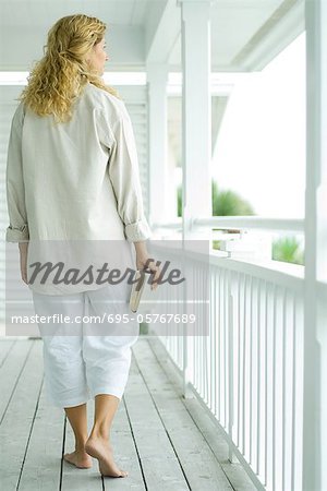 Woman walking on porch, carrying book, rear view