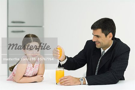 Father and daughter sitting together at table, man squeezing orange juice into glass, girl smiling at camera