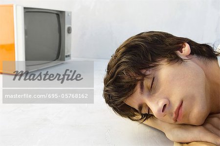 Young man resting head on arms, eyes closed, television in background