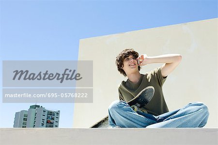 Teen boy using cell phone in urban setting, holding skateboard