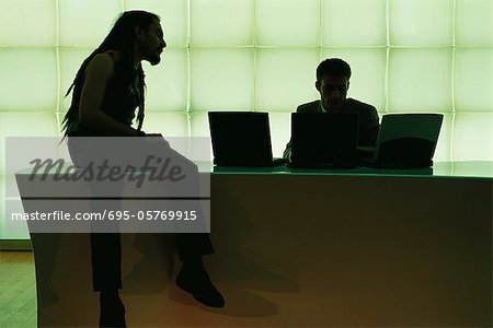 Man with dreadlocks sitting on desk speaking to businessman using laptop computer