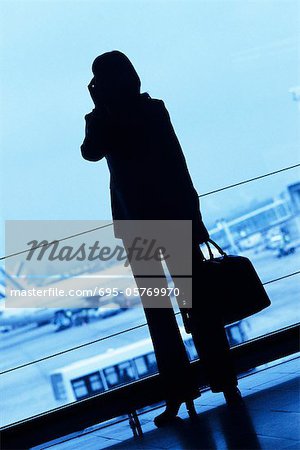 Silhouette of woman traveler using cell phone, holding travel bag in airport