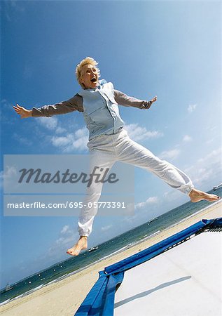 Mature woman jumping on trampoline at the beach, arms out, legs spread