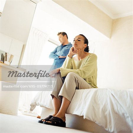 Man standing with arms crossed, behind woman sitting on edge of bed, low angle view